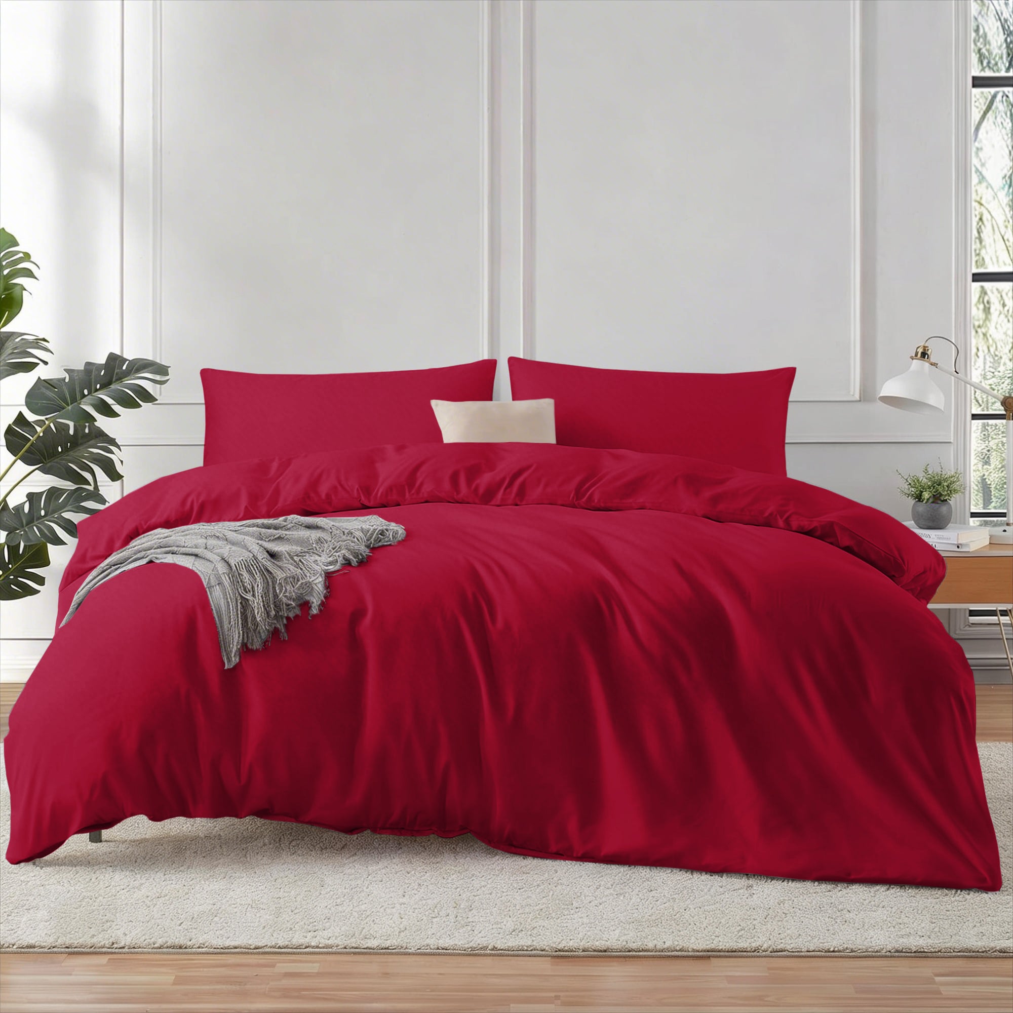 Red bedding set on a bed in a bedroom with a plant and window in the background.